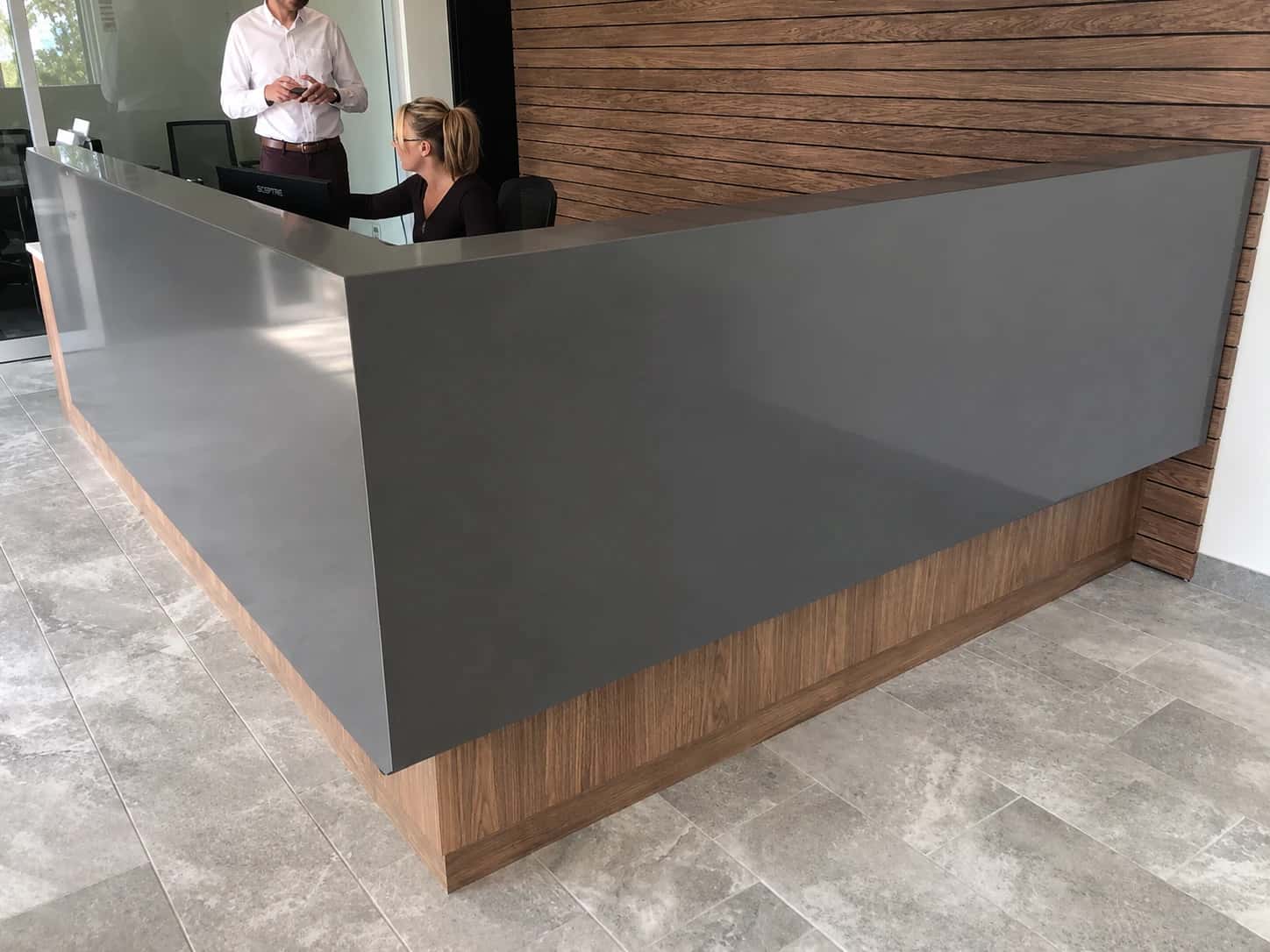 Completed coworking and flex office reception build-out by i30 Builders: an L-shaped gray quartz reception desk with a walnut base and a horizontal walnut slat feature wall, staffed at check-in.