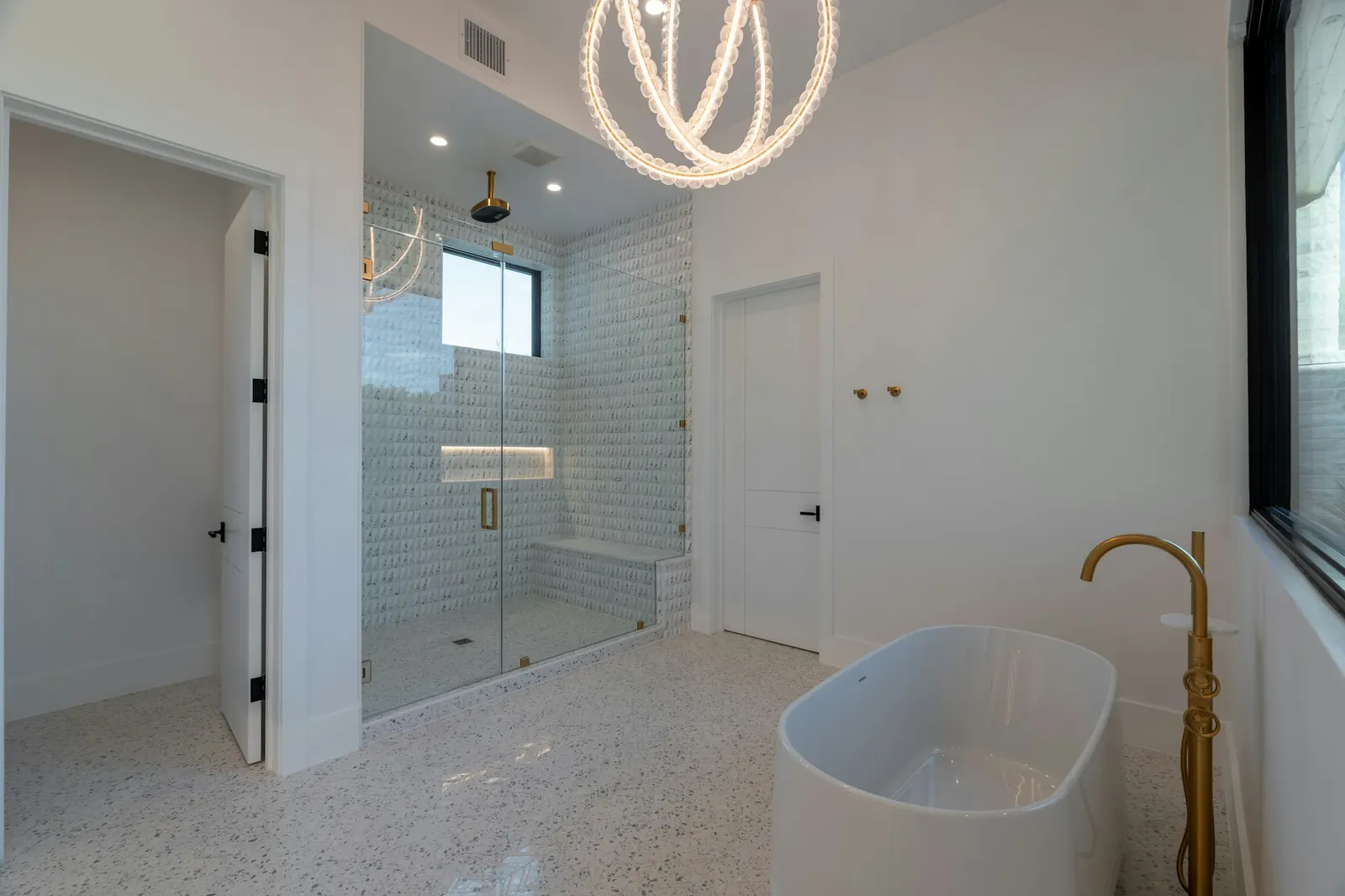 A large glass-enclosed shower with a bench and niche, and a freestanding tub with a floor-mounted brass filler.