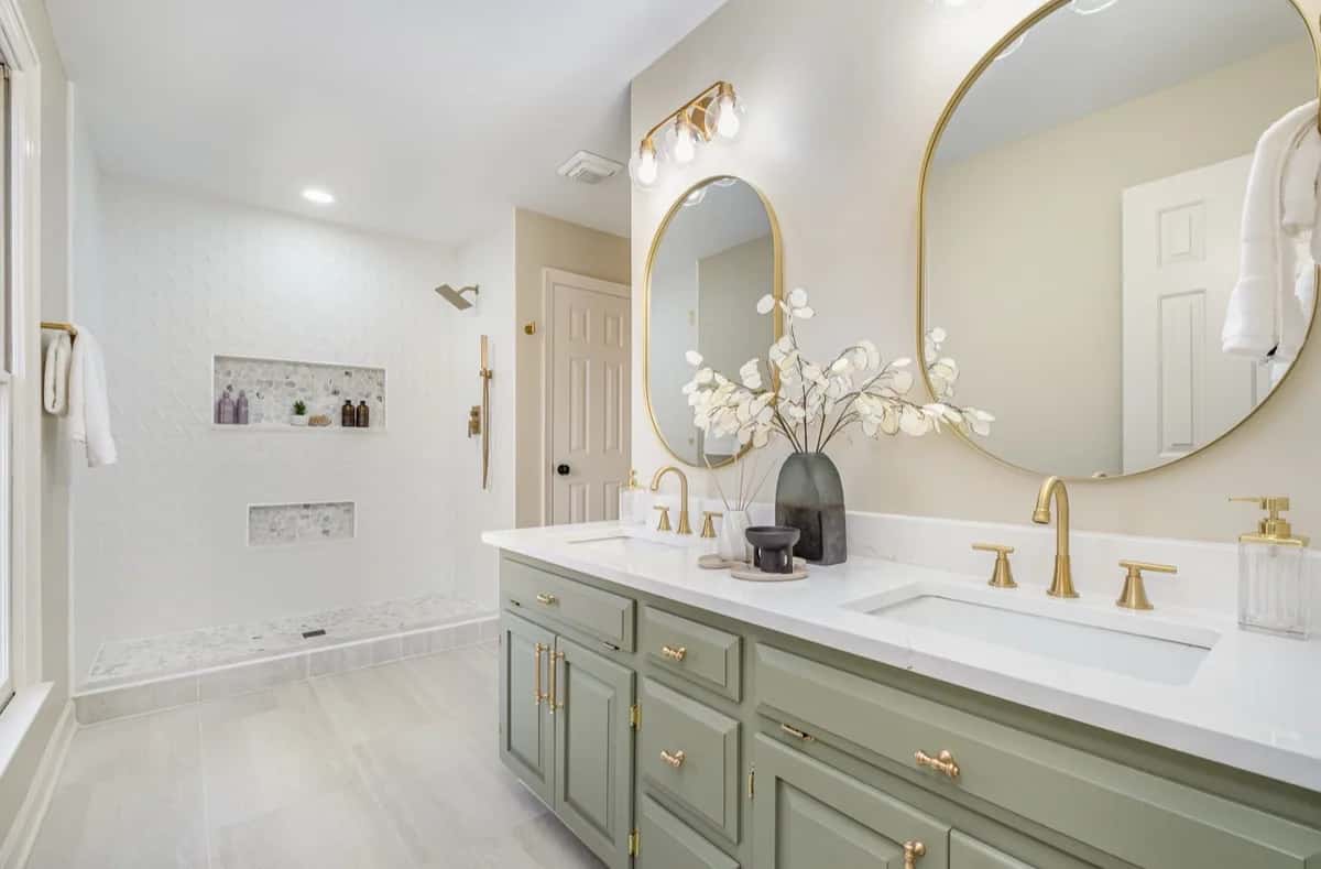 A remodeled bathroom with a sage double vanity, brass fixtures and oval mirrors, and a curbless walk-in shower with tiled niches.