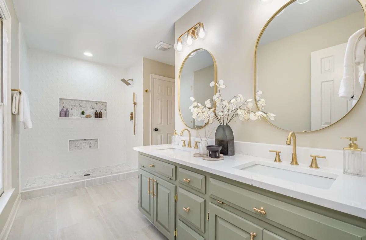 A curbless walk-in shower with tiled niches behind a sage double vanity with brass fixtures.