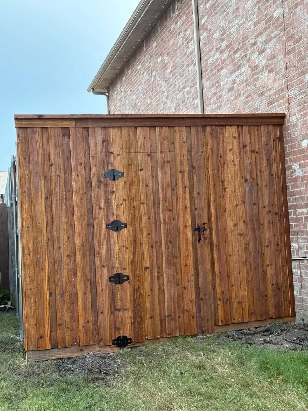 Board-on-board cedar enclosure against a brick house, with a top cap, black strap hinges and a thumb latch.
