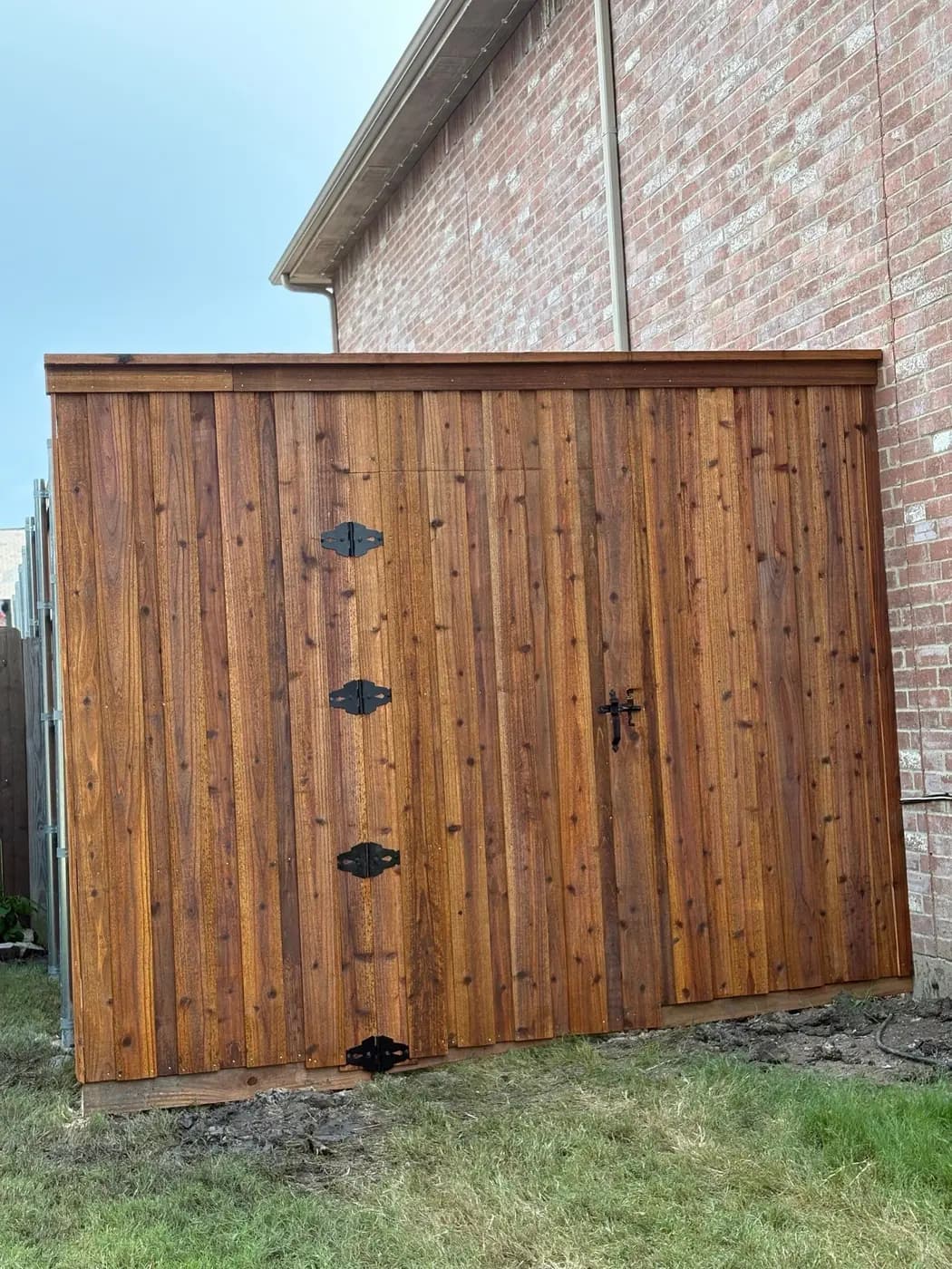 Board-on-board cedar enclosure against a brick house, with a top cap, black strap hinges and a thumb latch.