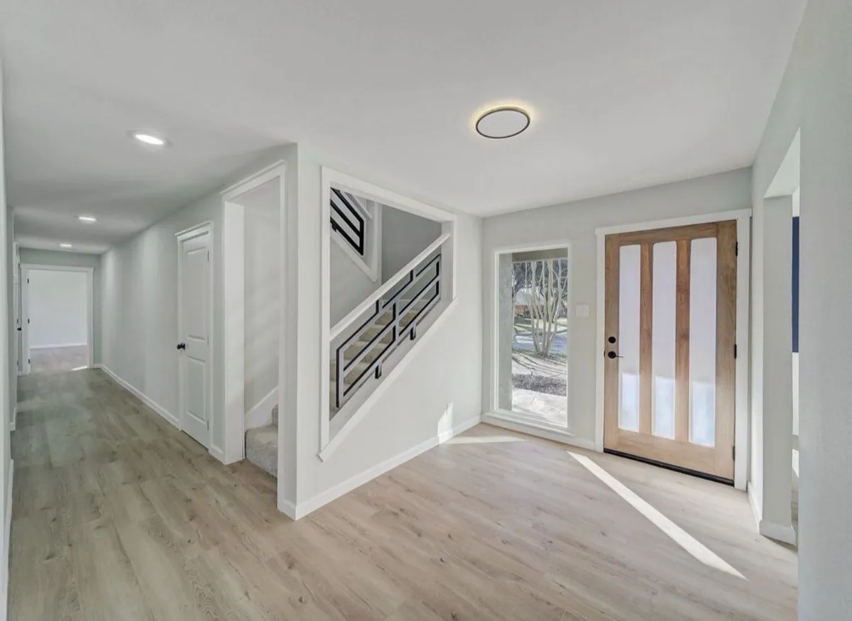 A remodeled entry and hallway with light plank flooring, a wood and glass front door with a sidelight, and a stair with a black geometric steel rail.