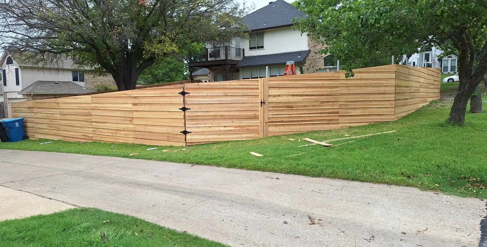 New unstained horizontal cedar fence stepping down a sloped yard beside a driveway, with a walk gate on black strap hinges.