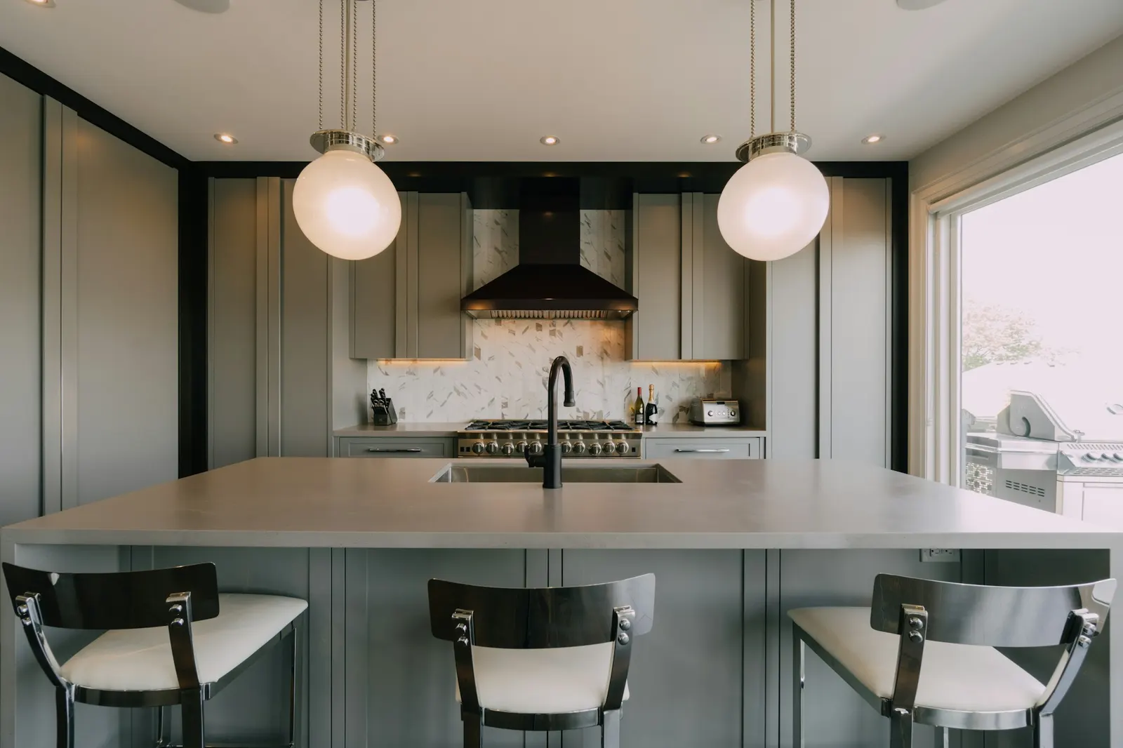 A kitchen with grey inset cabinetry to the ceiling, a professional gas range under a black hood, a marble mosaic backsplash, and a large island with seating under two globe pendants.