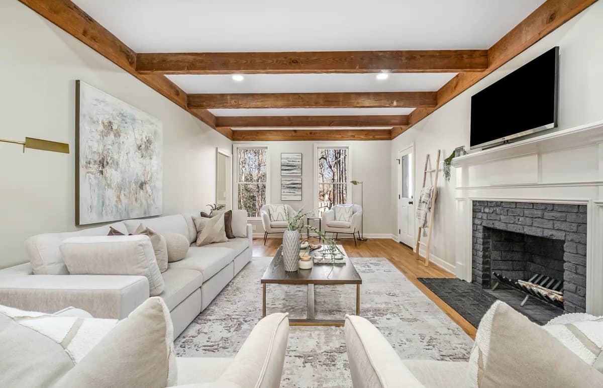 A living room with stained cedar ceiling beams, a white-painted brick fireplace with a white mantel and a TV above it, a hardwood floor and recessed lights.