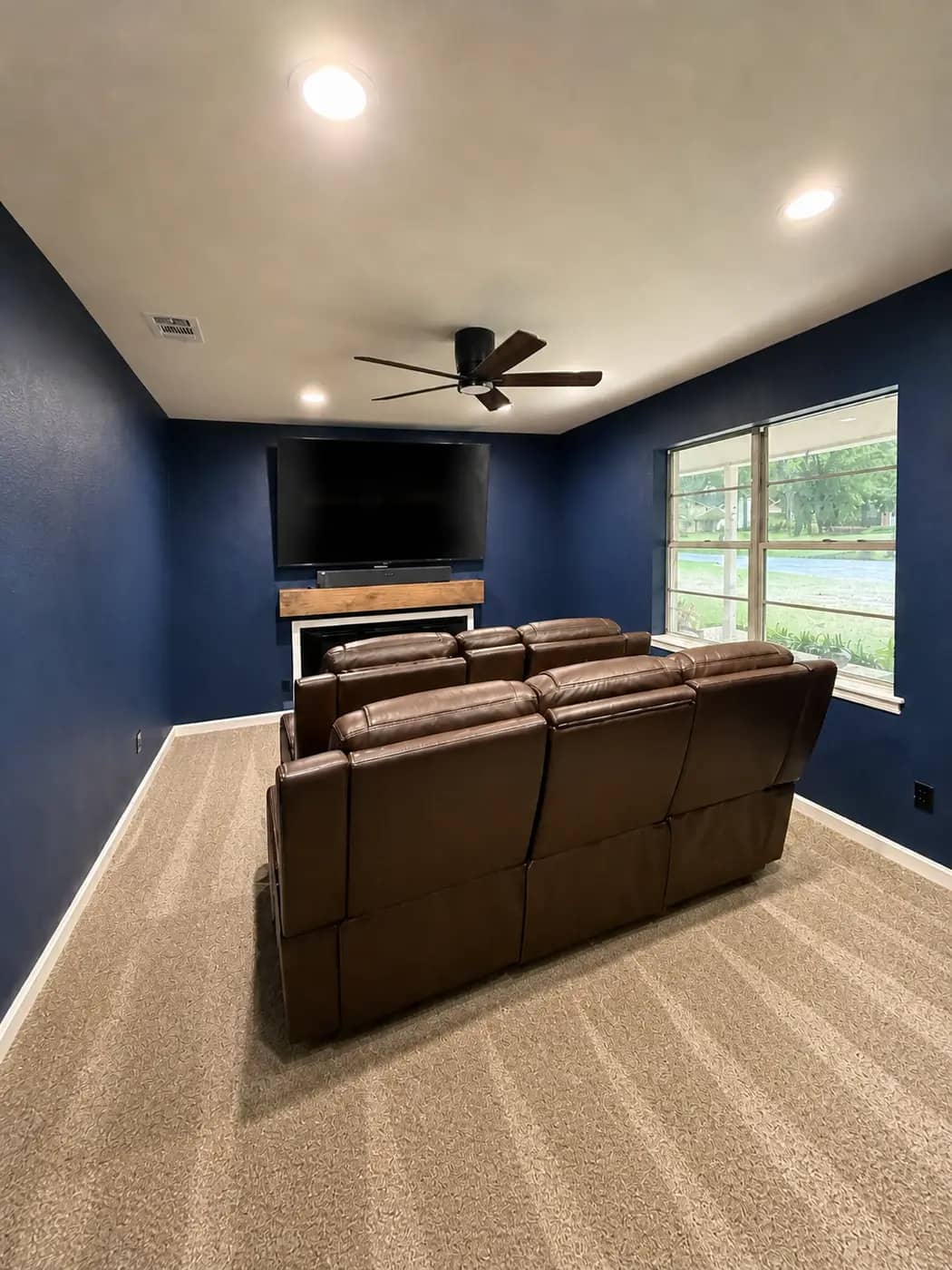 A living room in Longview remodeled into a media room: navy walls, a feature wall for the TV over the fireplace with a wood mantel, a new ceiling fan, recessed lights and carpet.