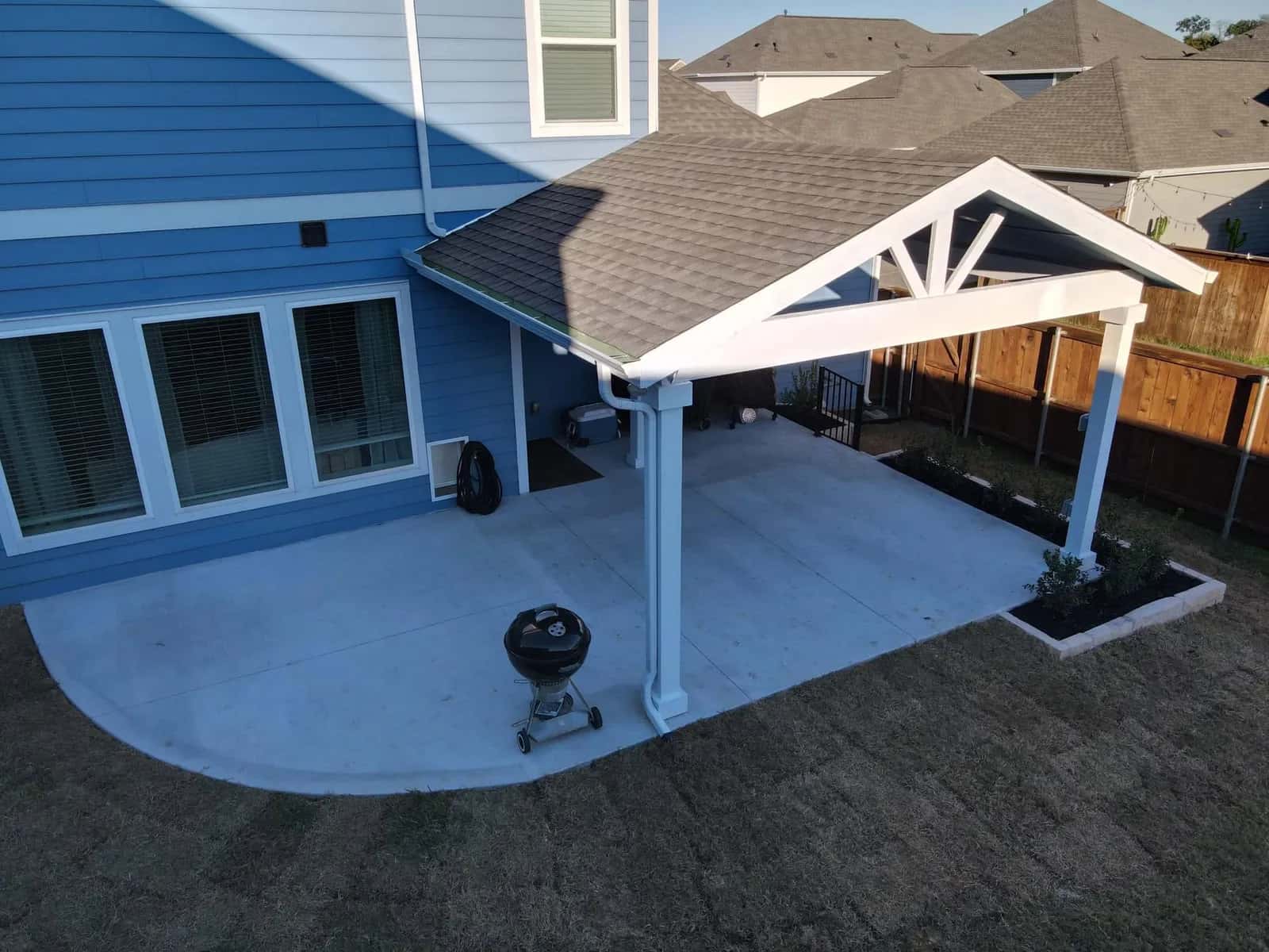 A new broom-finished concrete patio with a curved edge and steps, wrapping the back of a house under a white gabled patio cover, seen from above.