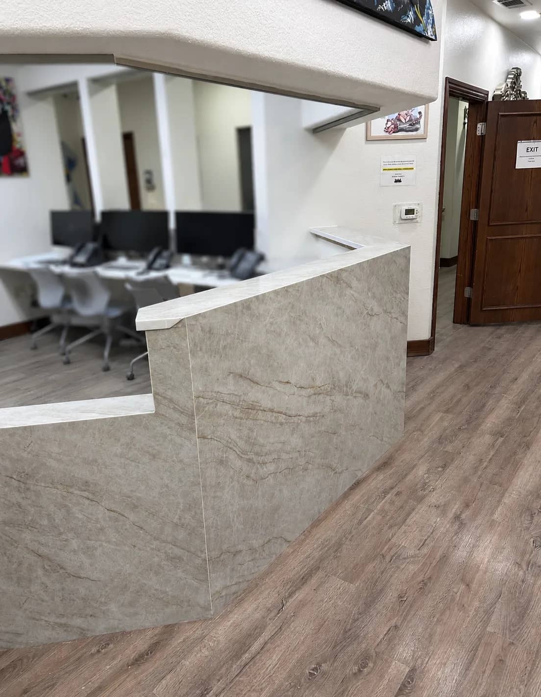 Finished medical office front desk clad in full-height quartz waterfall panels with mitered corners, over a wood-look plank floor, with the staff workstations behind it.