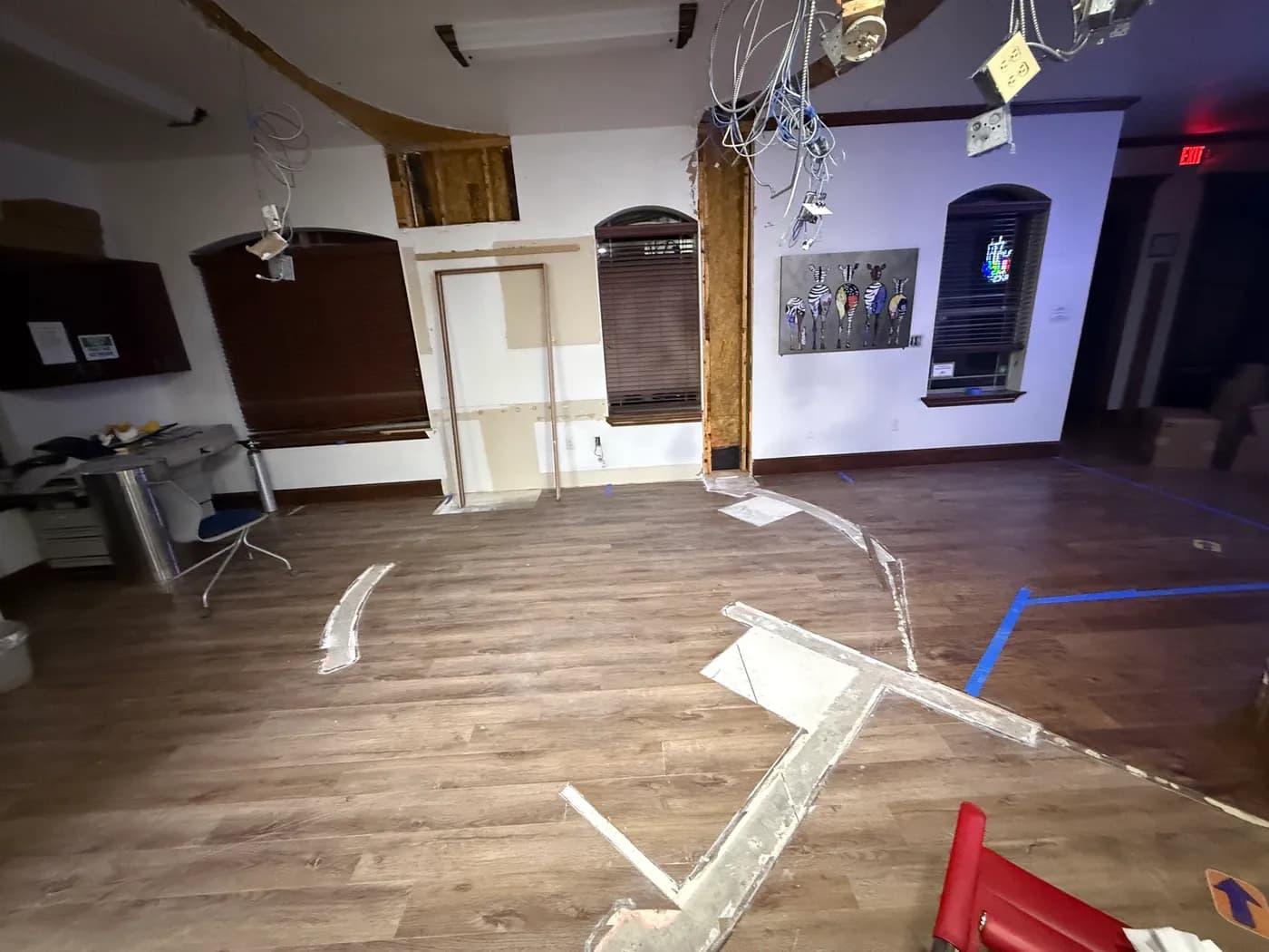 Pediatric clinic mid-demolition: ceiling opened up with conduit, junction boxes and data whips hanging, framing exposed at the column, and the new partition layout chalked and taped out on the floor.