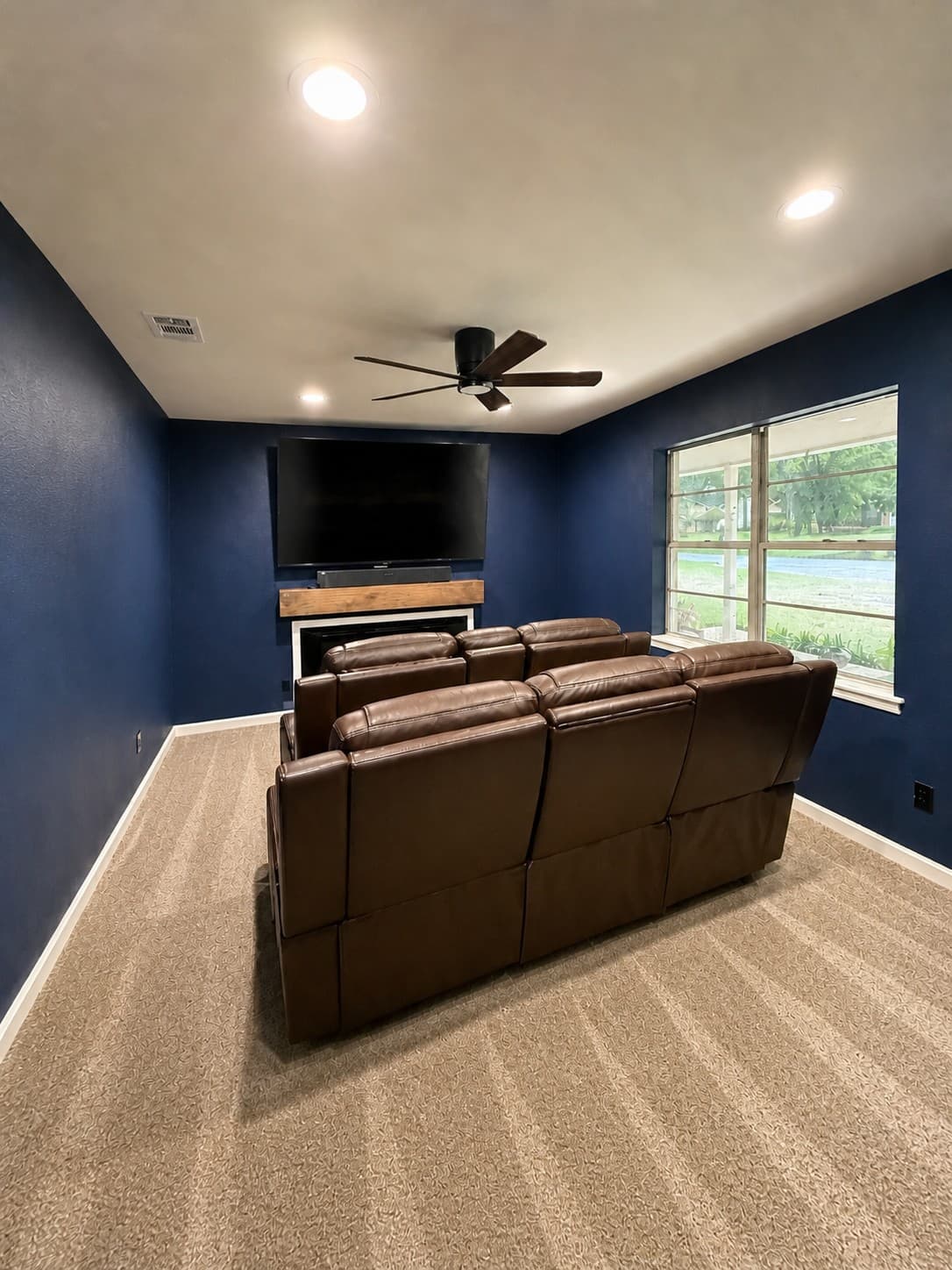 Completed media room — navy accent walls, mounted display with reclaimed-wood mantel, three-seat leather theater recliners, ceiling fan, and new striped low-pile carpet.
