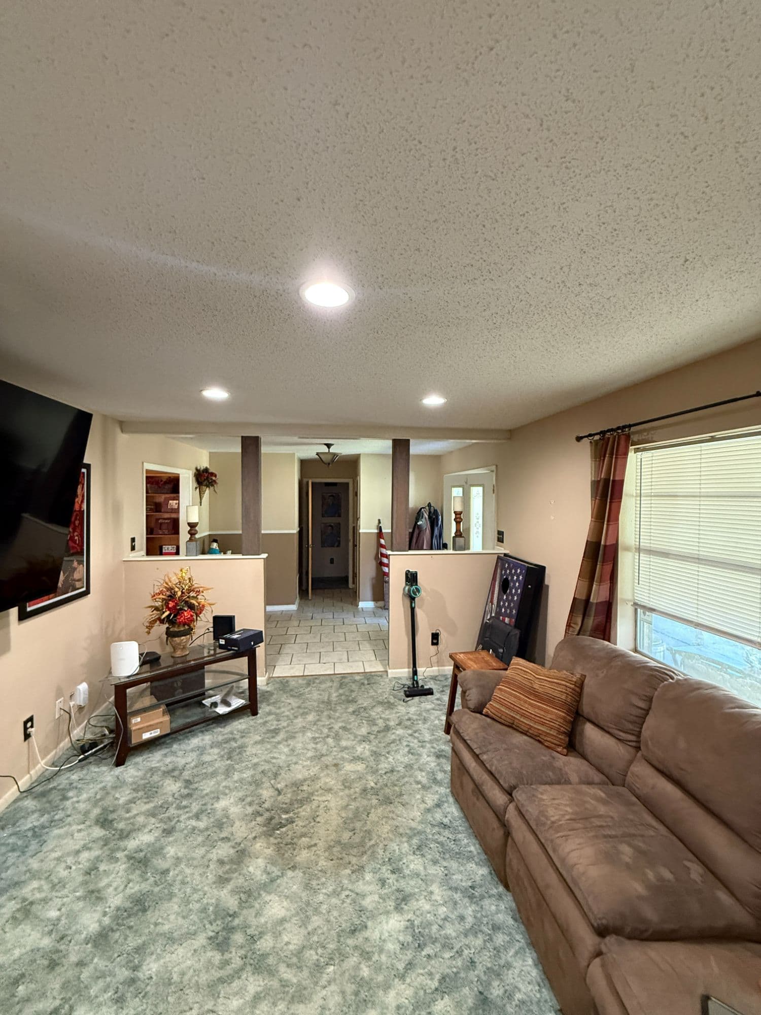 Living room before media-room conversion — beige walls, popcorn-textured ceiling, dated teal carpet, and mismatched furniture.