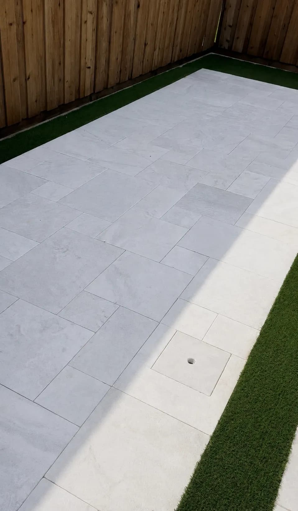 Light grey stone paver patio bordered with artificial turf against a cedar fence.